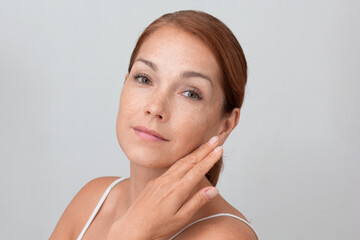 Portrait of caucasian middle aged woman face with freckles holding fingers on cheek, showing soft skin on white background looking at camera