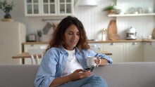 Young Spanish Woman Drinks Coffee And Writes SMS In Mobile Phone Sits On Sofa In Apartment. Satisfied Italian Female Smm Specialist Smiling Creates Post For Social Networks Makes Career As Freelancer