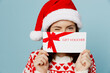 © ViDi Studio - Young merry woman wear red sweater Santa hat posing cover mouth gift certificate coupon voucher card for store isolated on plain pastel light blue cyan background. Happy New Year 2023 holiday concept.