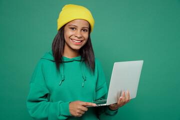 Wall Mural - Little smiling happy IT kid teen girl of African American ethnicity 13-14 years old wear casual hoody hat hold use work on laptop pc computer isolated on plain dark green background Childhood concept.