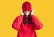 © Studio Romantic - Happy funny joyful smiling beautiful young woman in red sweatshirt puts warm red winter hat on her head and covers her eyes while standing on bright yellow studio background. Fashion concept