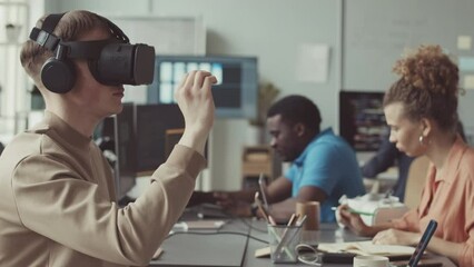 Young male software engineer in vr headset working with vr in modern office while his diverse colleagues coding on their computers