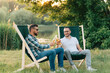 Two men sitting in camping chairs drinking beer and chatting outdoors.