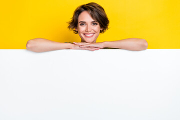 Wall Mural - Photo of cheerful pretty cute lady hides behind empty blank space billboard recommend beauty procedure isolated on yellow color background