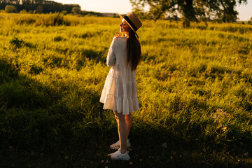 Wall Mural - back view of smiling redhead woman with closed eyes wearing straw hat and white dress standing posin
