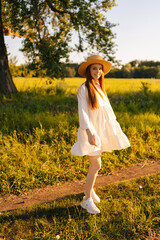Wall Mural - vertical portrait of smiling red-haired young woman in straw hat and white dress standing posing loo