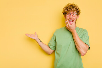 Wall Mural - Young caucasian man isolated on yellow background holds copy space on a palm, keep hand over cheek. Amazed and delighted.