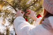 © Andriy Medvediuk - Child girl is decorating Christmas tree with shiny red balls outdoors before the holidays. Kid in warm jacket and hat is having fun outside in winter day. Merry Christmas and happy family holidays.