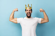 Portrait of excited strong confident man with beard wearing white T-shirt and gold crown, showing his biceps while raising arms, king of bodybuilding. Indoor studio shot isolated on blue background.