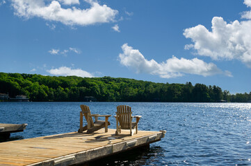 Two Adirondack chairs on a wooden dock facing a lake in Muskoka, Ontario Canada during a sunny summer morning. Cottages are nested between trees across the water.