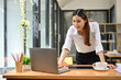 © bongkarn - Beautiful Asian businesswoman standing, leaning on table, looking at laptop screen