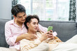 © TeTe Song - Cute Male Gay Couple Spend Time at Home. Young happy lgbtq couple Lying Down on a Sofa and Use a Smartphone, doing online shopping in ecommerce app together, having fun checking social media together