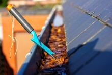 A Portrait Of A Roof Gutter Full Of Fallen Brown Autumn Leaves On A Sunny Day With A Small Blue Garden Shovel In It Of Someone Who Is Cleaning The Gutter During The Fall Season Annual Chore.