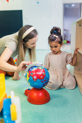 Wall Mural - blond teacher touching the little plastic globe and talking to a little girl at the nursery, vertical shot. High quality photo