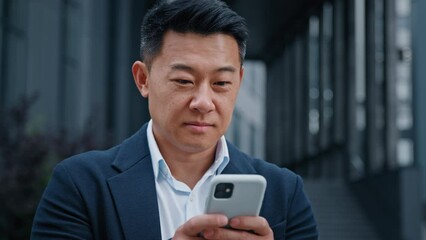 Close up Asian 40s businessman with smartphone texting browsing smiling looking at camera standing outdoors in city. Middle-aged man CEO has business chat using mobile phone check news in social net