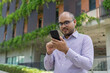 © Patcharanan - Bearded businessman smiling wearing glasses using smartphone device typing text message in social media stanting front modern building . Happy male spending free time in break of working.