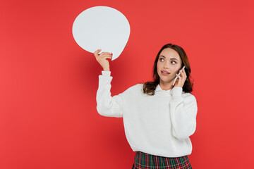 Wall Mural - brunette woman in sweater holding speech bubble and talking on smartphone on red background