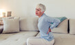 © Jelena Stanojkovic - Senior woman having back pain in the livingroom at home. Elderly woman suffering from backache at home. Unsmiling retired woman has a bad back on sofa.