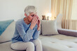 © Jelena Stanojkovic - Sad depressed woman at home sitting on the couch, looking down and touching her forehead, loneliness and pain concept. Depressed and sad grey hair mature woman sitting on bed at home during the day.