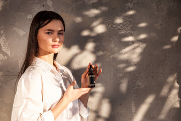 Wall Mural - Woman holding massage or cosmetics oil bottle for applying drops to skin or hair. Lady with oil at old wall background in foliage shadow. Concept of healthy lifestyle and self care. Copy text space