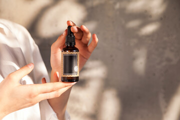 Wall Mural - Close up woman hands holding massage or cosmetics oil bottle for applying drops to skin or hair. Lady with oil at old wall background in shadow. Concept of healthy lifestyle and self care. Copy space