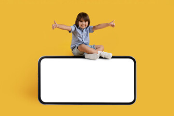 Cheerful little boy sitting on big mobile phone with empty screen and gesturing thumbs up, orange background