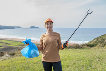 Positive Woman Standing On Hill With Trash Tongs And Sack