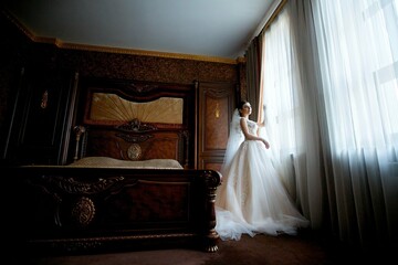 Beautiful young girl bride in a white wedding dress standing against the window.