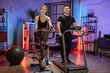 © sofiko14 - Beautiful smiling young woman doing cardio on stationary bike near her husband running on treadmill. Attractive male and female working out on fitness using exercise machines at evening time at home.