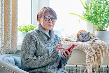 Wall Mural - Woman in sweater sitting in armchair near window with heating radiator and sleeping cat