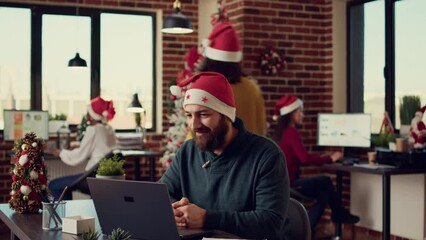 Wall Mural - Male employee talking on online videoconference call, wearing santa hat in festive office. Attending remote teleconference meeting during christmas season holiday, remote conference.