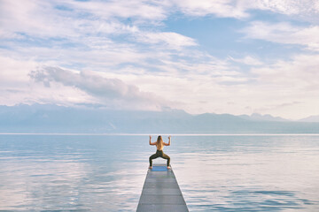 Outdoor portrait of young beautiful woman practicing yoga on dock by the lake, cactus pose, back view