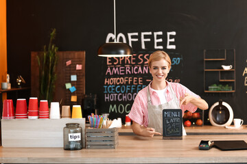 Wall Mural - Young female barista pointing at chalkboard with menu in cafe