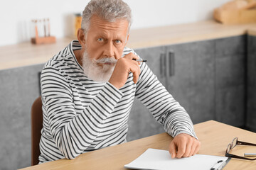 Wall Mural - Thoughtful mature man with pen and clipboard sitting at table in kitchen