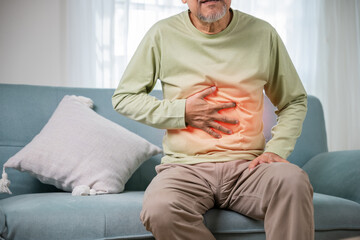 Senior Asian man sitting on sofa having suffering from stomach ache holding his stomach pain, Unhappy old aged man feeling discomfort because food poisoning, people health stomachache problem