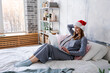 Happy girl in santa hat shopping online on laptop and sitting on bed next to christmas tree with lights in festive room in cozy clothes. Christmas sale and new year discount concept