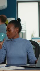 Wall Mural - African american woman taking a break from work using laptop to drink a sip of coffee or tea from cup in busy office. Casual startup employee working relaxed with statistics in business workplace.