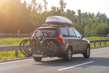 Bikes Fastened On Bicycle Holder Mounted On Back Side Of Car On Country Road. Brown Car With Roof Luggage Box And Trunk Bike Rack Driving On Highway.