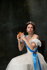 Wall Mural - Fast food or dieting. Young funny girl like queen or princess in white medieval outfit eating burger on dark background. Concept of comparison of eras, modern, fashion, beauty.