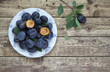 Fresh plums in bowl on wooden background with leaves and twig. Top view. Country style. Halved plum with seed as decoration. 