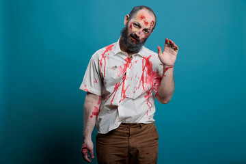 Fototapeta dangerous zombie waving hello at camera, doing salute gesture over blue background. creepy apocalyptic evil monster with bloody scars and wounds, brain eating aggressive corpse.