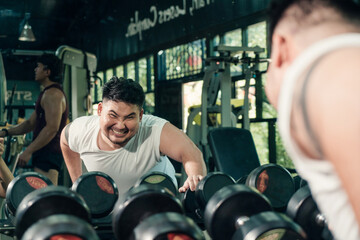 Asian plump man is intensely hevy weight lifting dumbbell in the gym while looking at the mirror, checking his gesture. Exercising weight training, Loss weight concept.