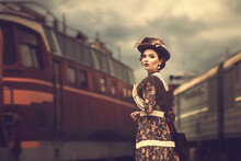 Beautiful Girl In A Historical Retro Dress On A Background Of An Old Steam Locomotive, Steampunk, At The Railway Station.