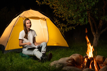 Female traveler is camping at a campsite. A young woman is sitting by the tent and reading a book in the camp by the campfire