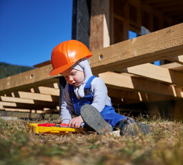 Wall Mural - Boy toddler playing as builder on construction site. Child carpenter in orange helmet and blue overalls learning to build wooden frame house outdoor on sunny day. Carpentry and workshop concept.