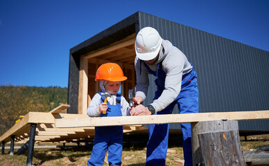 Wall Mural - Father with toddler son building wooden frame house. Male builders hammering nail into plank on construction site, wearing helmet and blue overalls on sunny day. Carpentry and family concept.