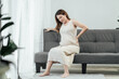 © ME Image - Full body of a beautiful pregnant asian woman is sitting on a couch in the living room, having a back pain as she carries the baby in her tummy. Pregnant problem.