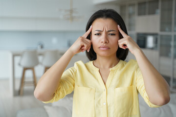 Frowning tired young woman suffering from headache, touches temples by fingers, standing at home