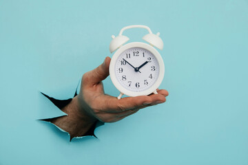 Hand holds alarm clock through a blue paper hole. Time management and deadline concept