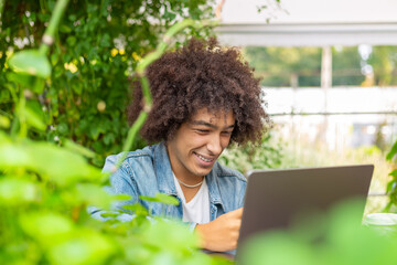 Smiling young mixed ethnic guy 20 years old, dressed in casual clothes, works on a laptop outdoors, in a green area in nature.Free Green open space for coworking, freelancer, relaxing, coffee, food. 